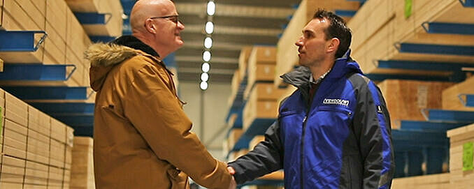 Zwei Personen schütteln sich die Hand. Im Hintergrund Kragarmregale.